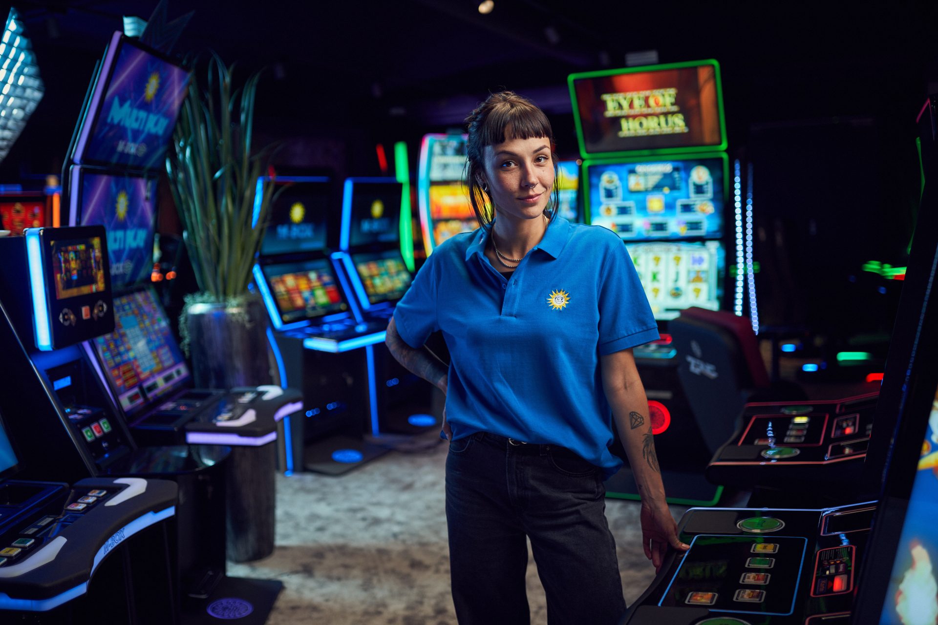 Frau, die an einem Spielautomaten lehnt und das MERKUR-Streetwear Polo Shirt in dunkelblau trägt