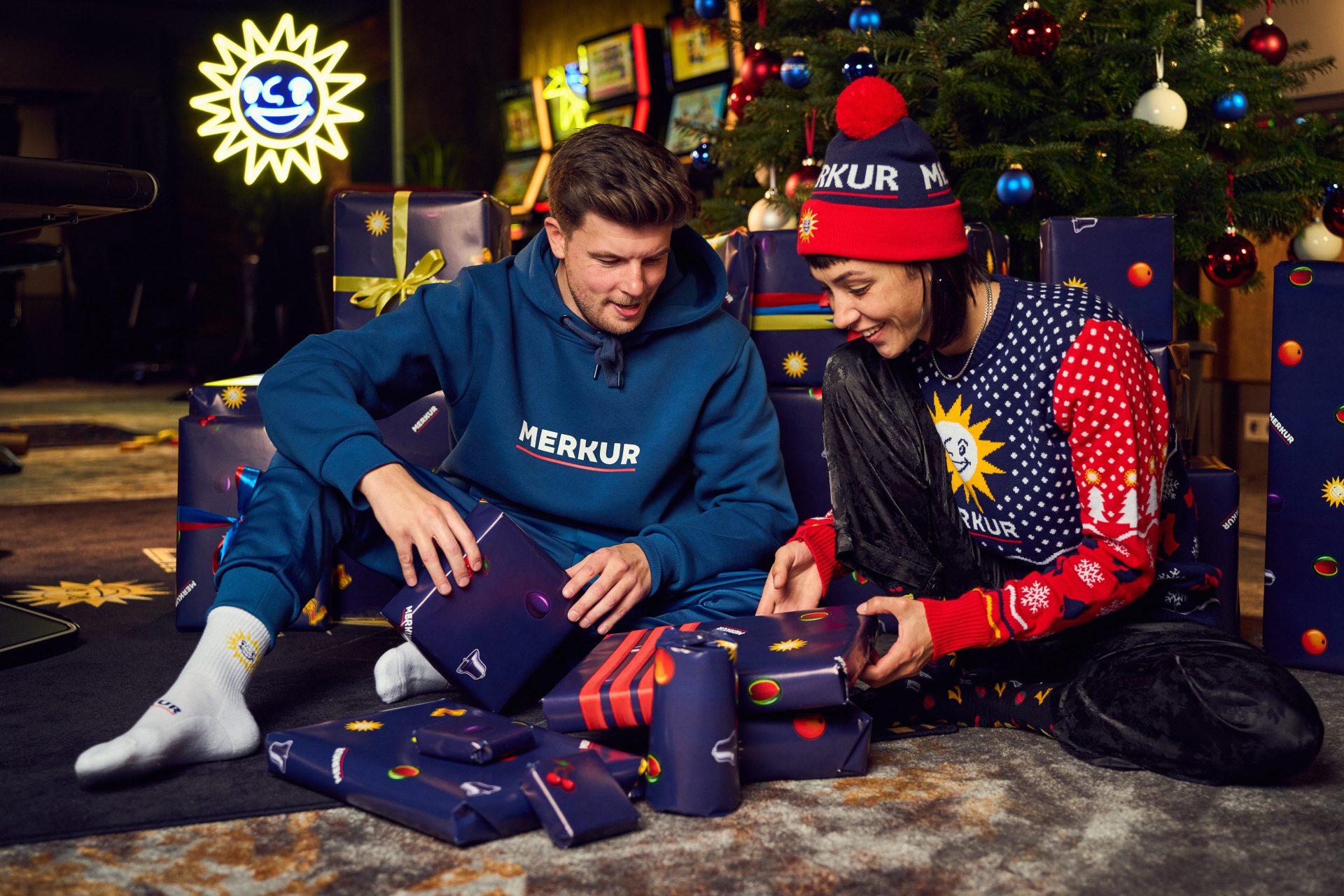 Zwei Personen, die unter einem geschmückten Tannenbaum sitzen und Geschenke auspacken. Dabei tragen sie MERKUR-Streetwear Kleidung