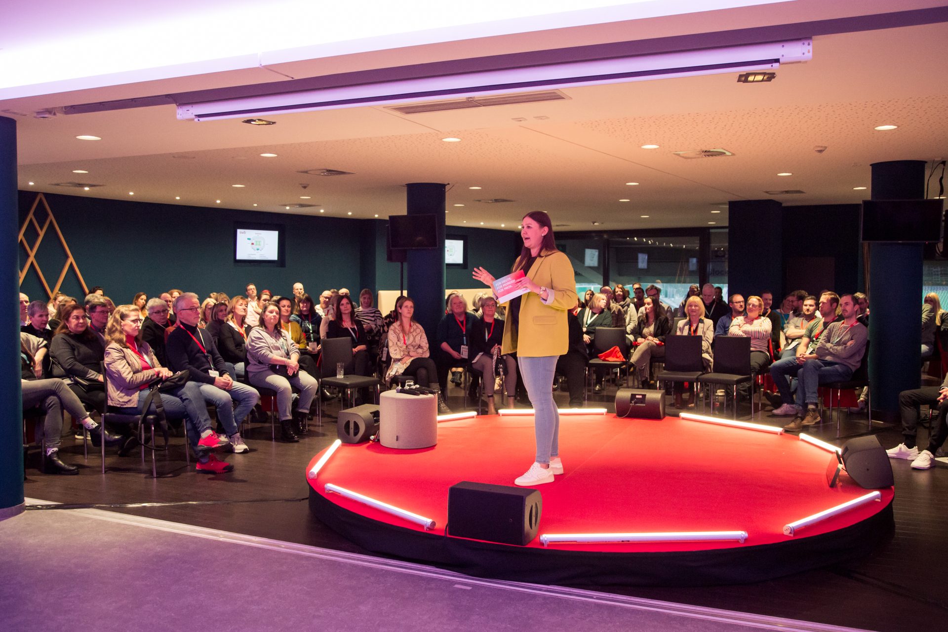 Mehrere Personen in Businesskleidung sitzen an Tischen in einem Konferenzraum und hören aufmerksam einem Vortrag zu.